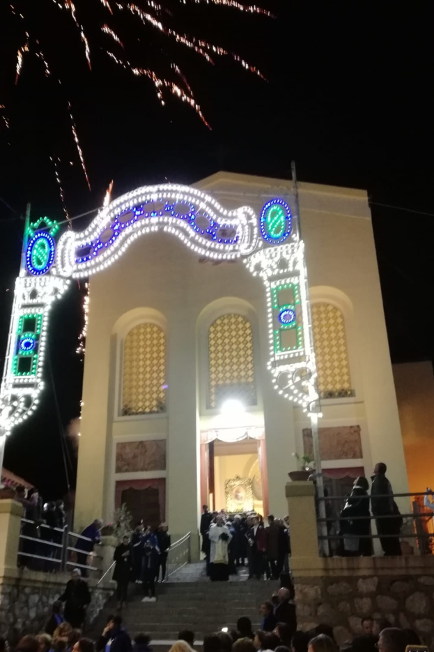 Eine Gruppe von Menschen steht vor einem Gebäude mit einem beleuchteten Torbogen, Treppen mit Geländern und Töpfen mit Pflanzen, während ein Feuerwerk den Himmel im Hintergrund erhellt.
