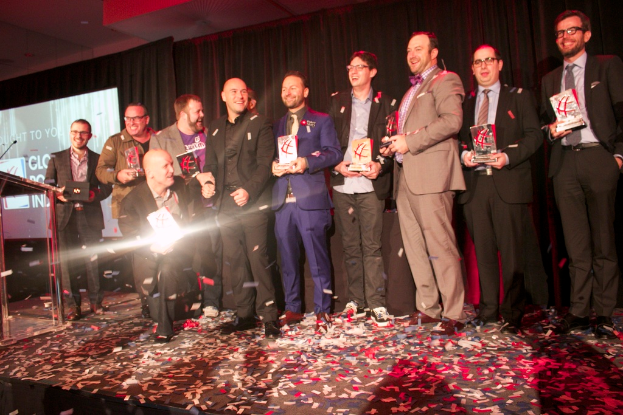 Eine Gruppe von Jungs steht auf einer Bühne und hält Schilde, mit einem Mann in der Mitte, der Licht reflektiert, einem Podium mit einem Mikrofon auf der linken Seite, einem Bildschirm und einem schwarzen Vorhang im Hintergrund sowie verstreuten Papierstücken auf der Bühne.