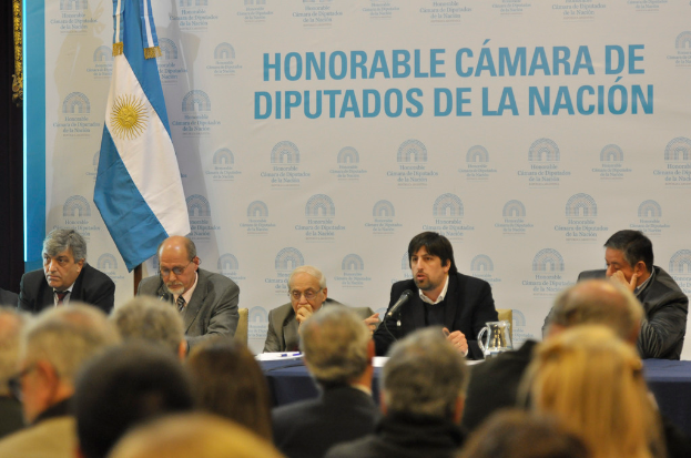 Eine Gruppe von Menschen sitzt an Tischen mit einem Krug, Papieren und einem Stift, während eine Person in der Mitte in ein Mikrofon spricht, mit einer weißen und blauen Flagge und einem Hintergrund, auf dem 'ehrenwerte kamara de' steht.