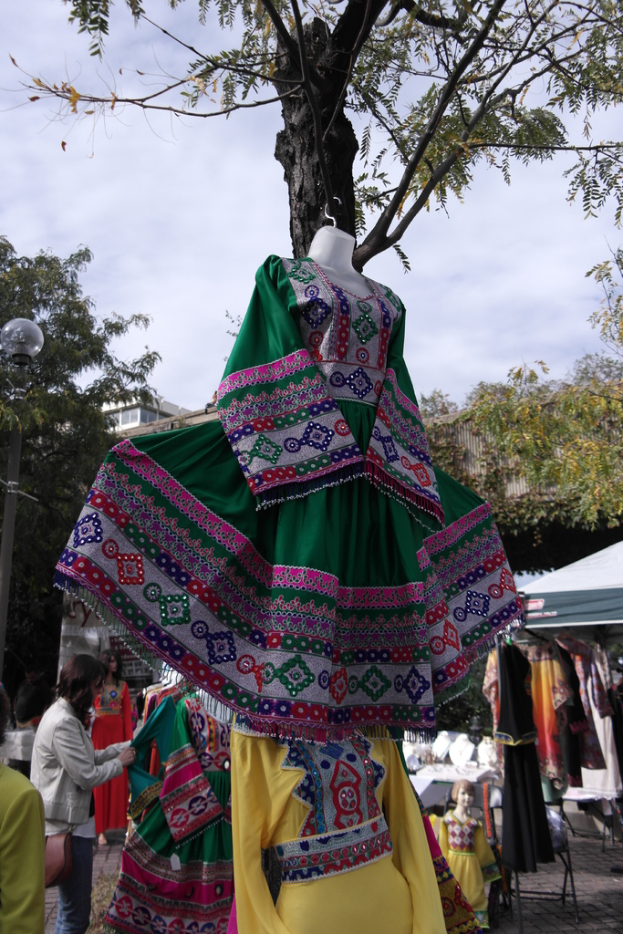 Eine Schaufensterpuppe steht in der Mitte eines belebten Einkaufsgebiets mit mehreren Geschäften, einer Person, die Kleider an einem Ständer arrangiert, einem Gebäude und Bäumen im Hintergrund.