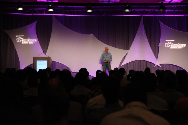 Ein Mann in einem blauen Hemd steht auf einer Bühne mit einem schwarz-weißen Vorhang und einem Metallrahmen mit Scheinwerfer und spricht zu einem sitzenden Publikum.