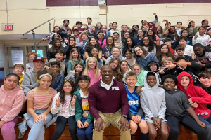 Gruppe von Schülern, die für ein Foto in einer Turnhalle posieren, wobei einige auf der Treppe sitzen und andere stehen, eine Tür und ein Ausgangsschild links sichtbar