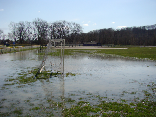 Ein Fußballtor steht in der Mitte eines überfluteten Feldes, umgeben von Gras, einem Zaun, Bäumen, Häusern und unter einem bewölkten Himmel.