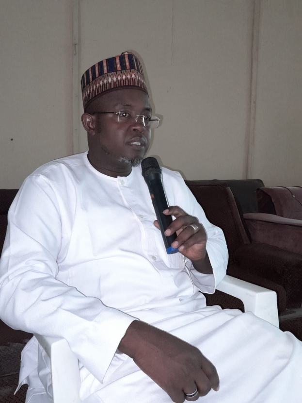 Ein Mann in einem weißen Kleid und einer Mütze sitzt auf einem Stuhl und spricht in ein Mikrofon in einem Raum mit zwei Sofas und einer Wand, auf der der Satz "Ich bin kein Lehrer, ich bin ein Lehrer." steht.