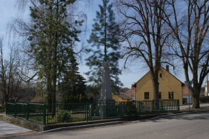 Denkmal für Holocaust-Opfer in der Mitte einer Straße, umgeben von einem Metallzaun, umgeben von Gras, Bäumen, Häusern, einem Straßenpfahl und einem bewölkten Himmel.