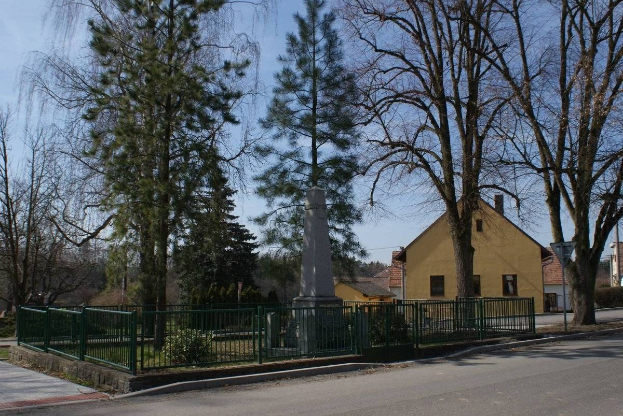 Denkmal für Holocaust-Opfer in der Mitte einer Straße, umgeben von einem Metallzaun, umgeben von Gras, Bäumen, Häusern, einem Straßenpfahl und einem bewölkten Himmel.