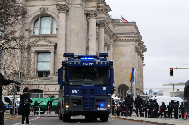 Eine Gruppe von Polizisten steht vor einem großen Gebäude mit Fenstern, Säulen und Bögen, mit Fahrzeugen auf der Straße, einer Person mit einer Kamera auf der linken Seite und einem klaren blauen Himmel mit Bäumen, Verkehrsampeln und Fahnen im Hintergrund.