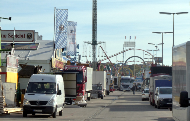Vollgestopfte Straße mit Lastwagen, Autos, Fußgängern, Laternen, Bannern, einem Turm, einem Vergnügungsgerät, Bäumen und einem bewölkten Himmel.