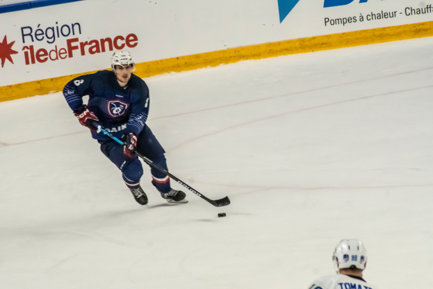 Ein Hockey-Spieler in Uniform und Helm hält einen Stock auf dem Eis, mit einer textbedeckten Wand im Hintergrund.