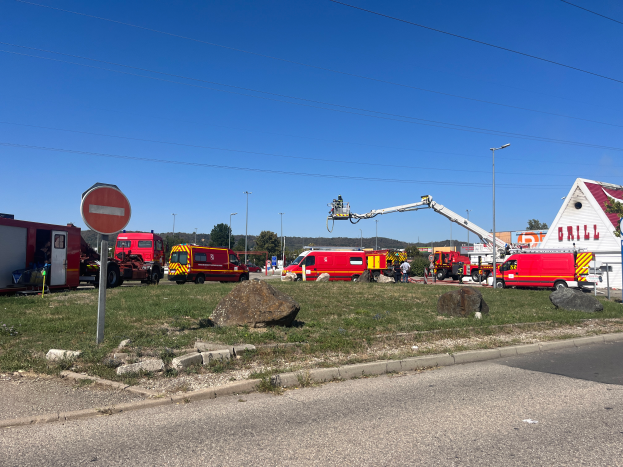 Eine Gruppe von Feuerwehrautos steht an der Seite einer Straße, umgeben von Gras, Steinen, einem Schild, einem Haus, Masten, einem Kran, Drähten, Bäumen und einem bewölktem Himmel, wahrscheinlich nach einem Brand an einer Tankstelle.