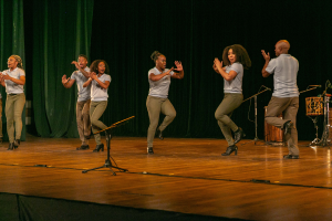 Eine Gruppe von Menschen auf einer Bühne mit Mikrofonen, Musikinstrumenten und einem Vorhang im Hintergrund, die Hände klappend, als würden sie feiern