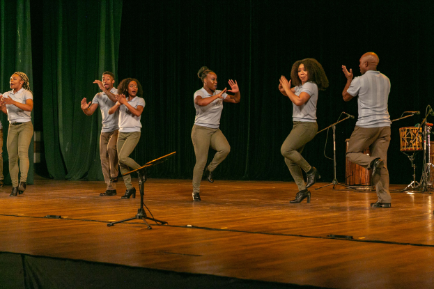Eine Gruppe von Menschen auf einer Bühne mit Mikrofonen, Musikinstrumenten und einem Vorhang im Hintergrund, die Hände klappend, als würden sie feiern