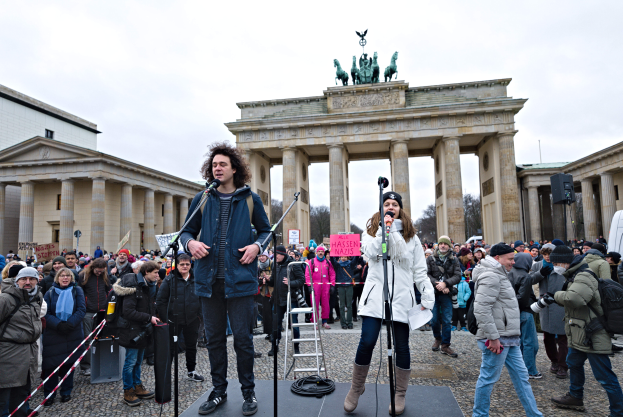 Eine Gruppe von Menschen auf einer Bühne vor dem Brandenburger Tor in Berlin, mit zwei Personen, die Mikrofone halten, und einer weiteren Person mit einer Kamera, sowie einer Leiter im Hintergrund und Gebäuden, Bäumen und einem klaren blauen Himmel.