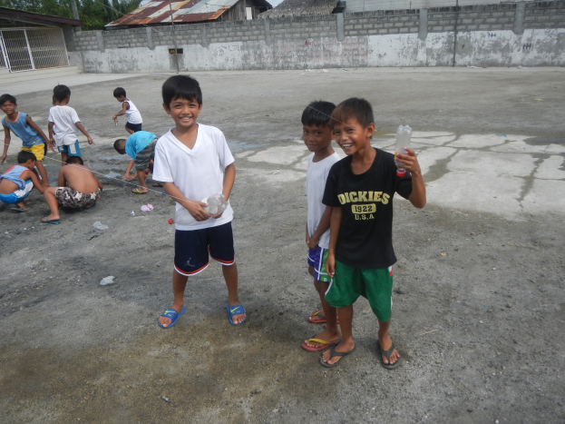Eine Gruppe junger Jungs, die zusammen in einer Parkplatz stehen, einige halten Flaschen, mit Häusern, Bäumen, Pfählen und einem klaren blauen Himmel im Hintergrund.