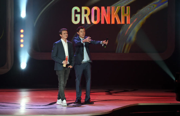 Eine Person in der Mitte mit einem Mikrofon haltend und sprechend, mit einer anderen Person links stehend und lachend, vor einem Bildschirm mit Text und Lichtern.