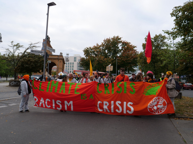 Eine Gruppe von Menschen marschiert eine von Bäumen gesäumte Straße entlang und hält ein Banner mit der Aufschrift "Klima-Krise ist eine Krise", mit Fahrzeugen und Gebäuden im Hintergrund unter einem klaren blauen Himmel.