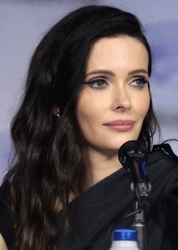 Krysten Ritter auf der Comic Con in San Diego, in einem schwarzen Kleid, vor einem Mikrofon mit einer Flasche unten, mit einem unscharfen, nebeligen Hintergrund.