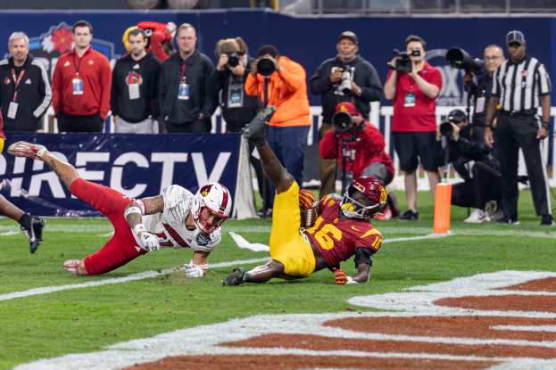 Ein Fußballspieler taucht in die Endzone ein, um einen Touchdown zu erzielen, mit einer Gruppe von Menschen am Boden, einige halten Kameras, und eine Fahne mit Text im Vordergrund, und Geländer, Pfosten und andere Gegenstände im Hintergrund.