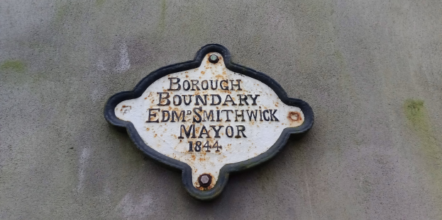 Metallschild an der Seite eines Gebäudes, das die Borough-Grenze markiert, mit "Borough-Grenze Edm Smithwick Bürgermeister" in fetten weißen Buchstaben auf einem schwarzen Hintergrund.
