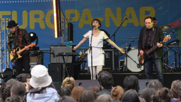 Vier Menschen, die auf einer Bühne Musik machen, mit einer Frau in einem weißen Kleid, die singt, und zwei Männern, die Gitarren spielen, während das Publikum darunter eine Wand mit Lautsprechern im Hintergrund sieht.