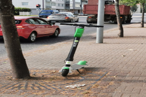 Elektrischer Roller auf der Straße neben einem Baum geparkt mit Fahrzeugen und Gebäuden im Hintergrund.