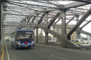 Ein blauer Bus fährt über eine Brücke mit Autos, umgeben von Gebäuden, Bäumen, Polen und einem klaren blauen Himmel.