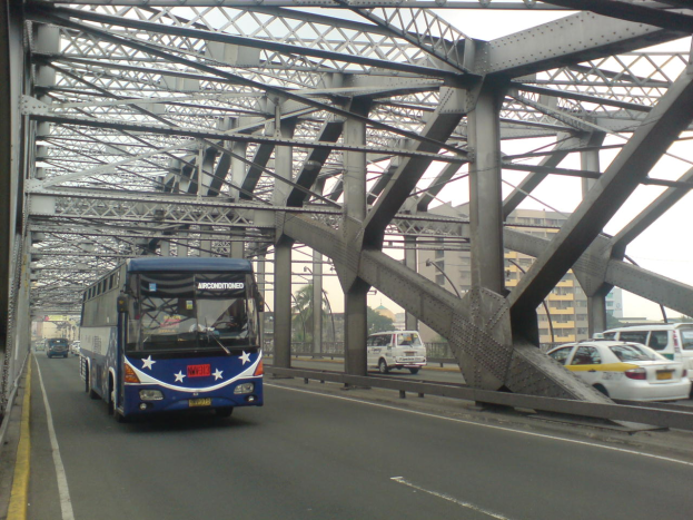 Ein blauer Bus fährt über eine Brücke mit Autos, umgeben von Gebäuden, Bäumen, Polen und einem klaren blauen Himmel.