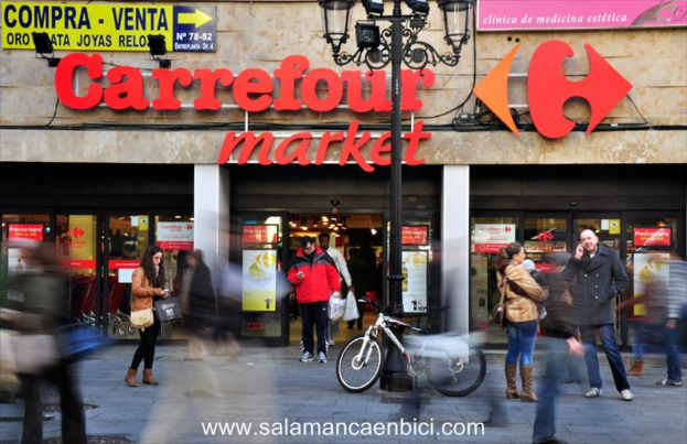 Ein belebter Einkaufsmarkt mit Menschen vor einem Einkaufszentrum, ein schwarzer Mast mit Lampen, ein Fahrrad neben dem Mast und ein Richtungsschild an der Wand.