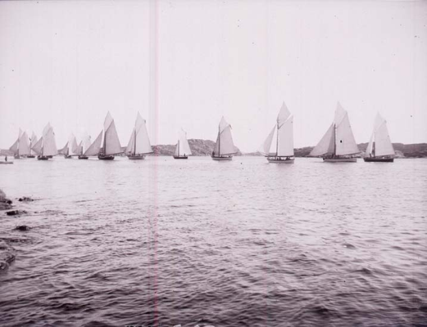 Schwarzes und weißes Foto von Segelbooten auf dem Wasser mit Bergen im Hintergrund, klarem Himmel darüber und 'New York Yacht Club'-Text unten.