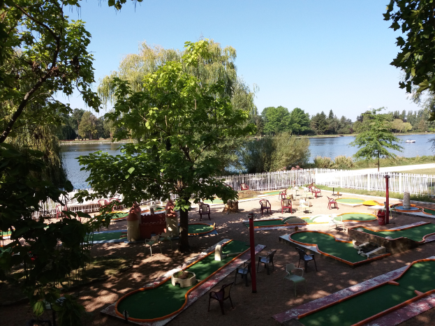Ein Minigolfplatz in einem Park mit grünem Gras, Bäumen und einem Zaun, mit Stühlen und Tischen, einem Gewässer mit einem Boot und einem sichtbaren Himmel.