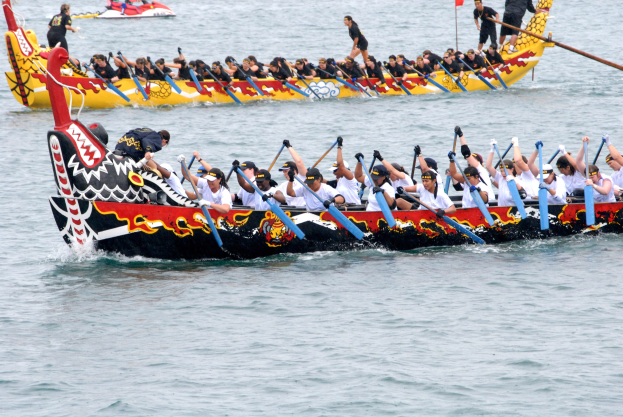 Gruppe von Menschen in einem Drachenboot, das auf dem Wasser rennt, einige halten Paddel, mit einem anderen Boot und einer Flagge im Hintergrund.