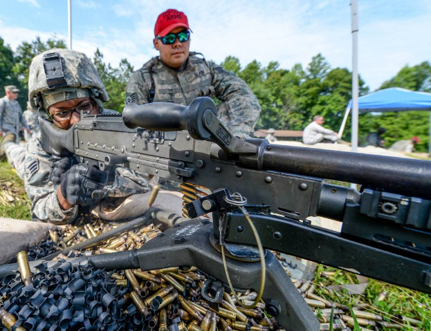 Eine Person auf einem Armee-Übungsplatz schießt mit einer Waffe, während daneben eine Person mit roter Mütze steht. Im Hintergrund sind Bäume, Zelte und Menschen zu sehen, unter einem klaren, sonnigen Himmel, mit Patronenhülsen und Gras im Vordergrund.