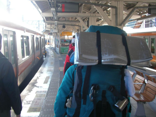 Ein Bahnhofsgebäude mit Menschen neben einem Zug, einem Stand in der Mitte, einem weiteren Zug auf der rechten Seite und einer Bank in der rechten unteren Ecke.