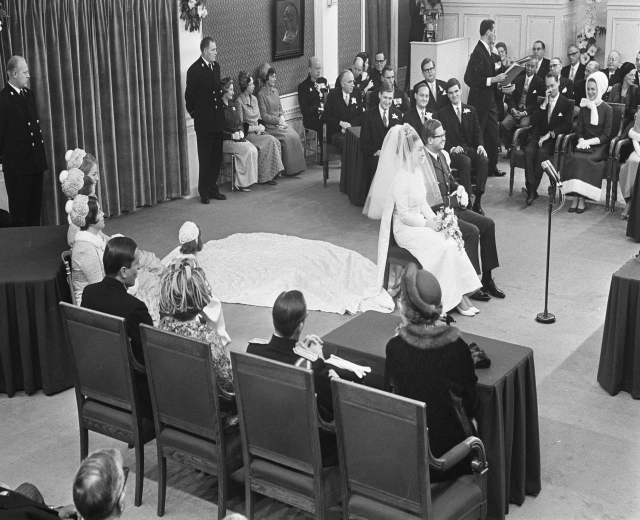 Schwarz-weiß-Foto einer Hochzeitszeremonie mit sitzenden und stehenden Menschen, einem Mann mit Mikrofon, einer Frau mit Blumen, einem Podium mit Mikrofon, Blumenschmuck, einem Vorhang, einem gerahmten Foto an der Wand und einer Tür im Hintergrund.