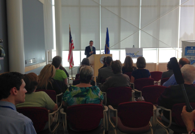 Ein Mann steht an einem Rednerpult und spricht zu einem Publikum, das auf Stühlen sitzt, mit zwei Fahnen daneben und einer Wand, Säulen und Fenstern im Hintergrund.