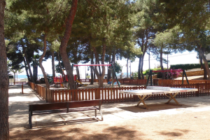 Ein Park mit einem zentralen Tischtennis-Tisch, Bänken, Schaukeln, Pfosten, Pflanzen und Bäumen, vor einer Wasserfläche und einem klaren blauen Himmel.