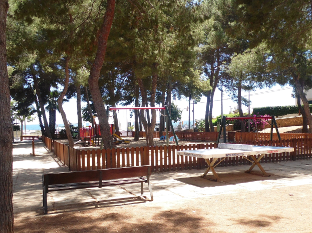 Ein Park mit einem zentralen Tischtennis-Tisch, Bänken, Schaukeln, Pfosten, Pflanzen und Bäumen, vor einer Wasserfläche und einem klaren blauen Himmel.