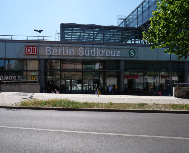 Großes gläsernes Berliner Südkreuz-Bahnhof mit sichtbarem Text, umgeben von Straßeninfrastruktur, Fahrzeugen, Fußgängern, Fahrrädern, Bäumen und einem klaren Himmel.
