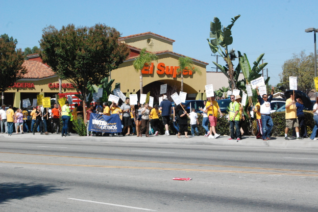 Eine Gruppe von Menschen mit Schildern marschiert eine Straße entlang, gesäumt von Bäumen, Büschen und Gebäuden mit Namensschildern, mit Autos im Hintergrund.