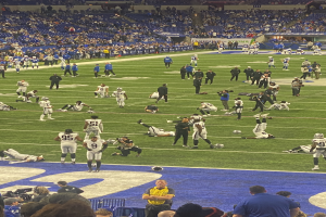 Ein Fußballspiel wird in einem Stadion gespielt, während Menschen auf dem Rasen stehen und einige auf Stühlen im Hintergrund sitzen und eine Person eine Kamera hält, was auf eine Live-Übertragung hinweist.