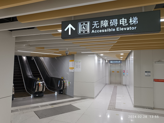 Rolltreppe links mit einer 'Barrierefreier Aufzug' Tafel darüber, zwei Türen rechts, Deckenleuchten und Plakate an der Hintergrundwand.