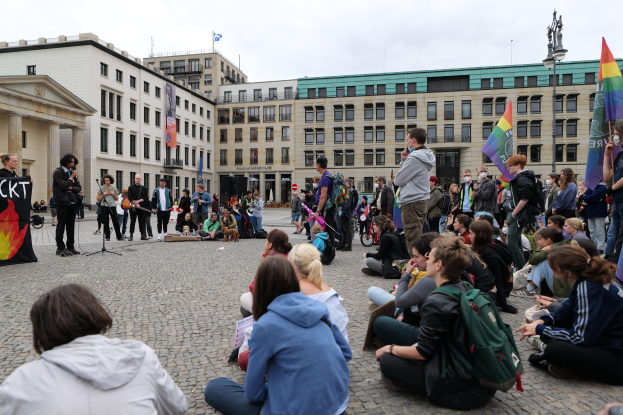 Eine Gruppe von Menschen, die auf dem Boden vor einer Menge sitzen, die Fahnen und Transparente schwingt, bei einer Demonstration in Berlin, mit einem Redner, einem Denkmal und Gebäuden im Hintergrund.