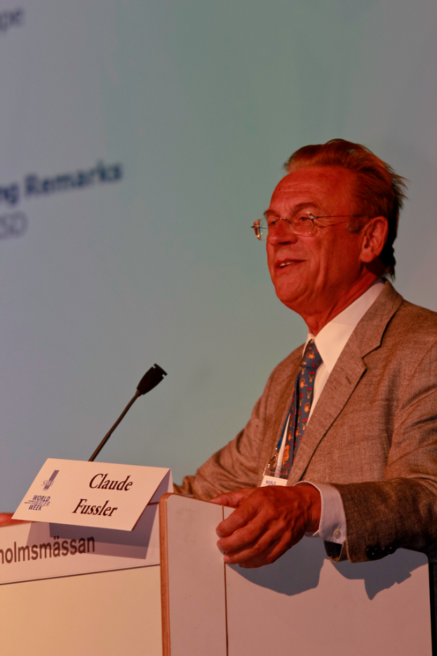 Ein Mann in einem weißen Hemd und einem grauen Blazer steht lächelnd vor einem Podium mit Mikrofon und Namensschildern, im Hintergrund ein Projektionsschirm oder eine Fahne mit Text.