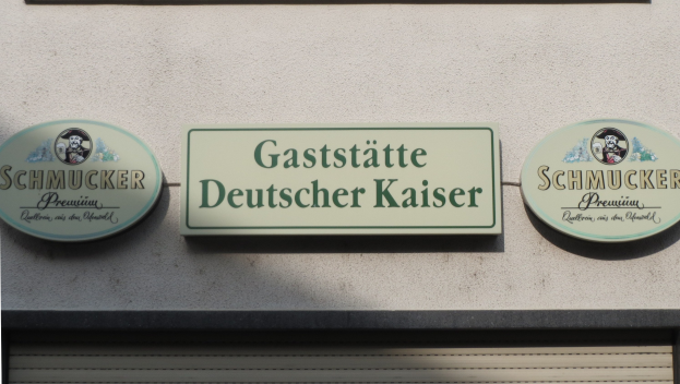 Drei hölzerne Bretter mit der Aufschrift "Gaststätte Deutscher Kaiser Schmucker" sind an einer Gebäudewand über einem geschlossenen Rollladen angebracht.