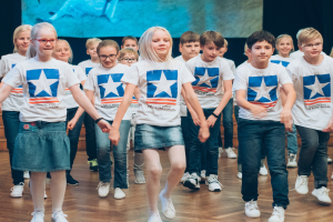 Gruppe von Kindern in weißen Hemden und blauen Shorts, die auf einem Holzboden tanzen, während im Hintergrund ein Bildschirm zu sehen ist.