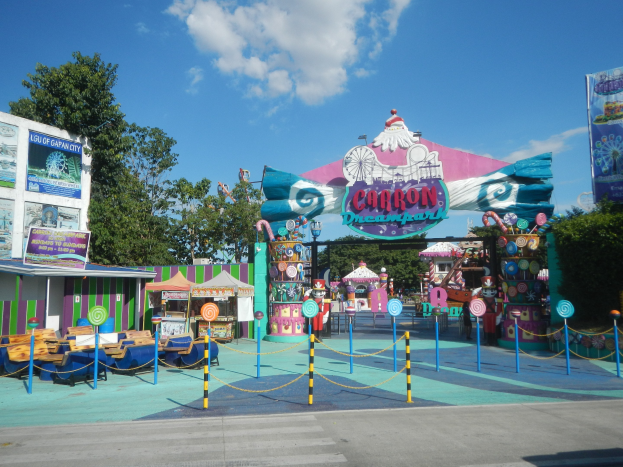 Karussell am Parkeingang mit Straße, Zaun, Buden, Schildern, Bäumen und bewölktem Himmel im Hintergrund.