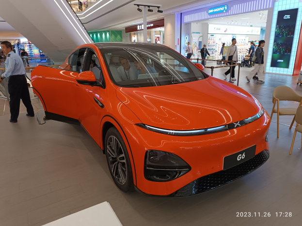 Ein oranges Elektroauto in einer Ausstellungshalle umgeben von Menschen, mit Stühlen und einem Wagen rechts und Geschäften im Hintergrund.