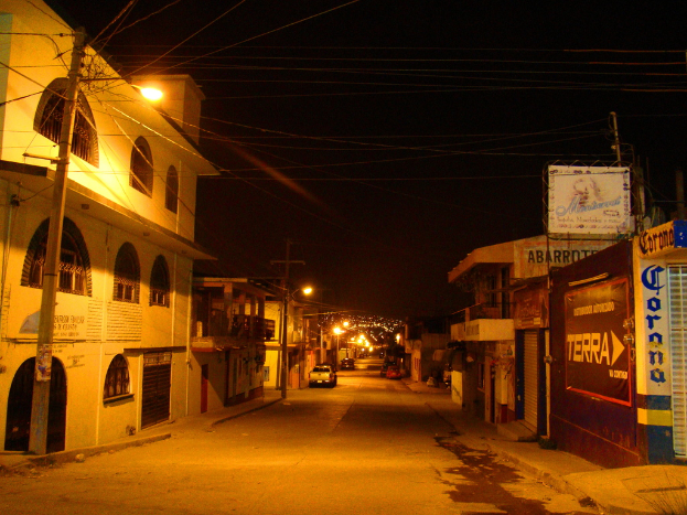 Eine StraÃŸenszene mit GebÃ¤uden und StrommÃ¤sten auf beiden Seiten, parkenden Fahrzeugen vor den GebÃ¤uden und einer StraÃŸe in der Mitte.