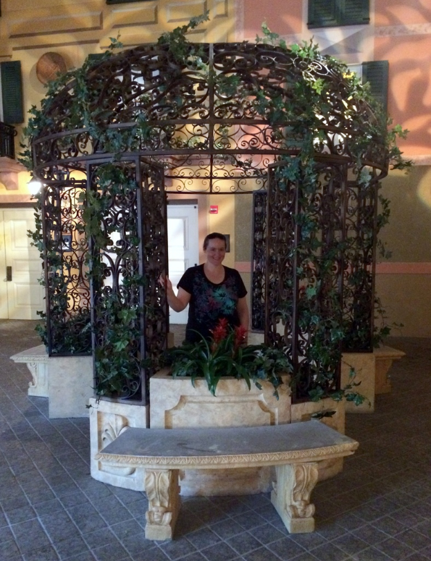 Eine Bank im Vordergrund mit einer verzierten Eisen Säule und Kletterpflanzen dahinter, mit einer Frau in einem schwarzen Kleid und einem Gebäude mit Wänden, Fenstern und Türen im Hintergrund.