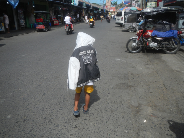 Ein junger Junge geht eine Straße entlang, die von Motorrädern und anderen Fahrzeugen sowie Gebäuden mit Schildern gesäumt ist, unter einem klaren blauen Himmel mit Bäumen und Pfählen im Hintergrund.